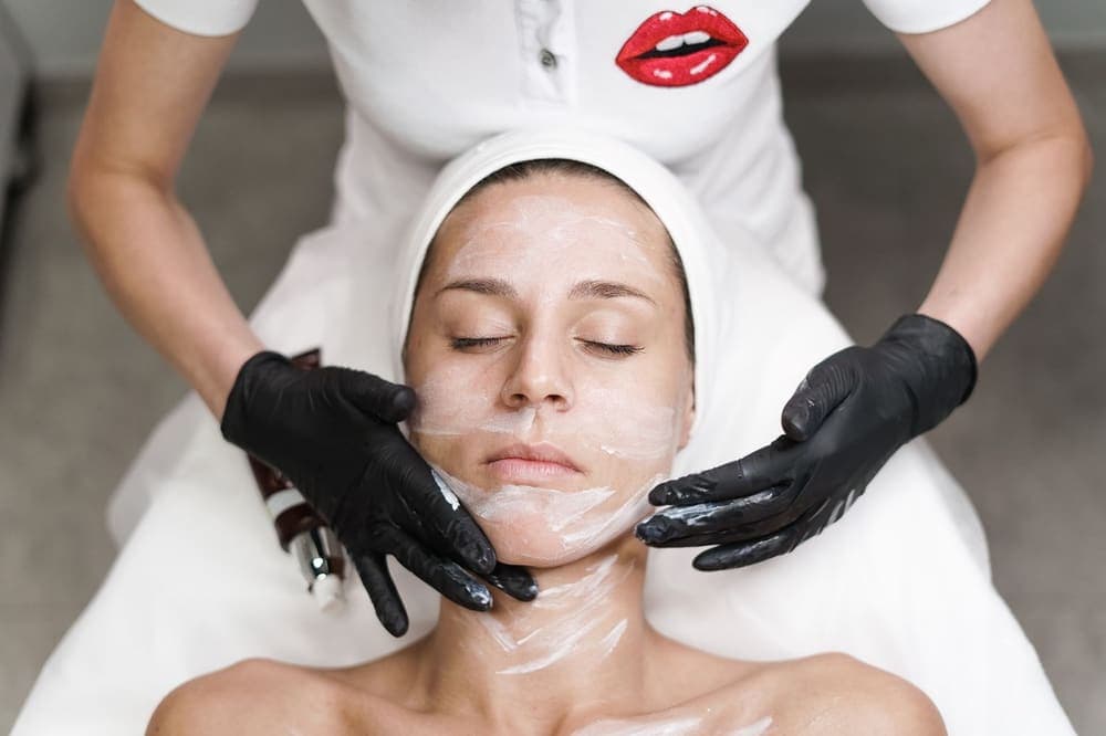 An esthetician in black gloves smoothing a white mask over a relaxed client's face