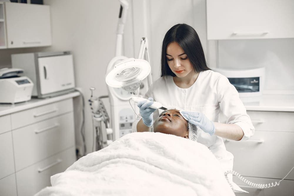 An esthetician using a facial device on a client lying under a white blanket